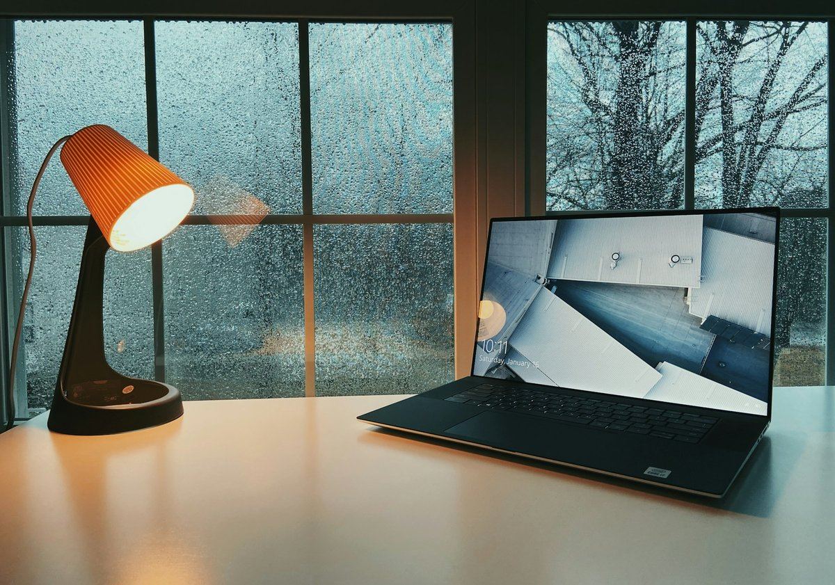 Lampe à lumière orangée allumée près d'un ordinateur portable, devant une fenêtre couverte de gouttes de pluie