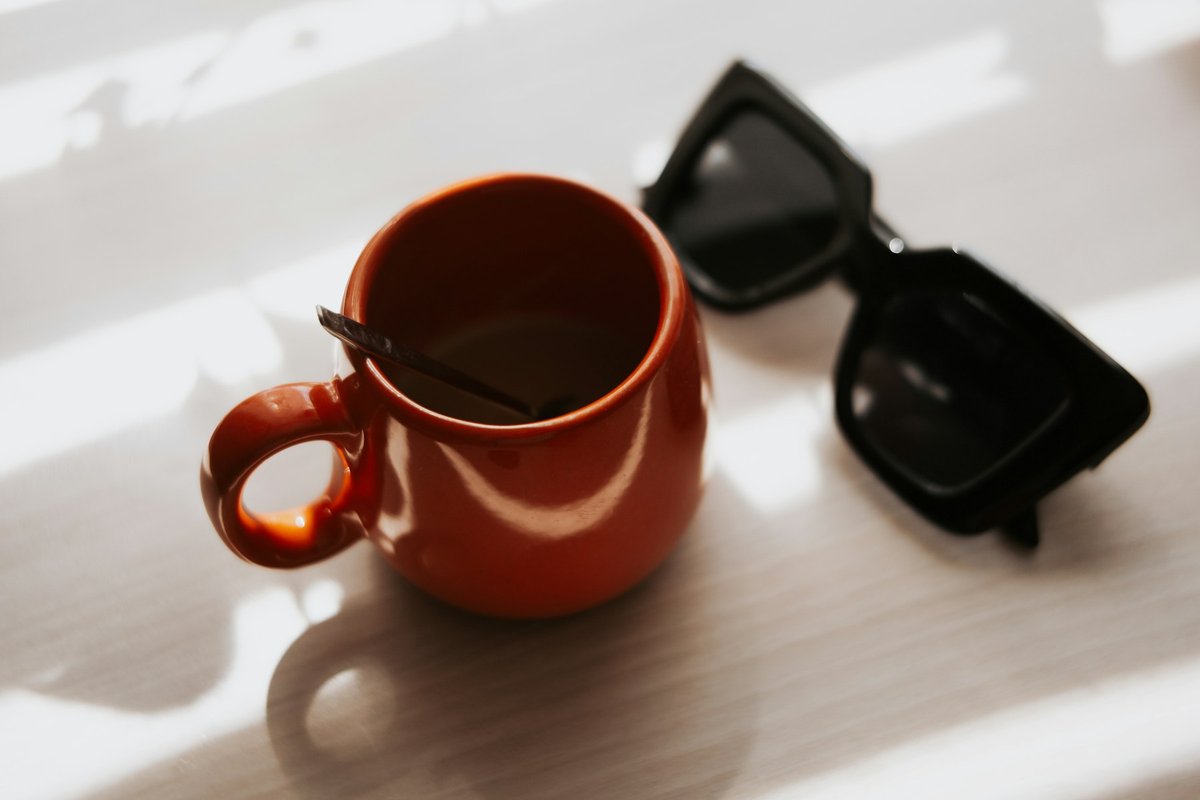 Lunettes de soleil noires posées à côté d'une tasse orange sur une table blanche ensoleillée